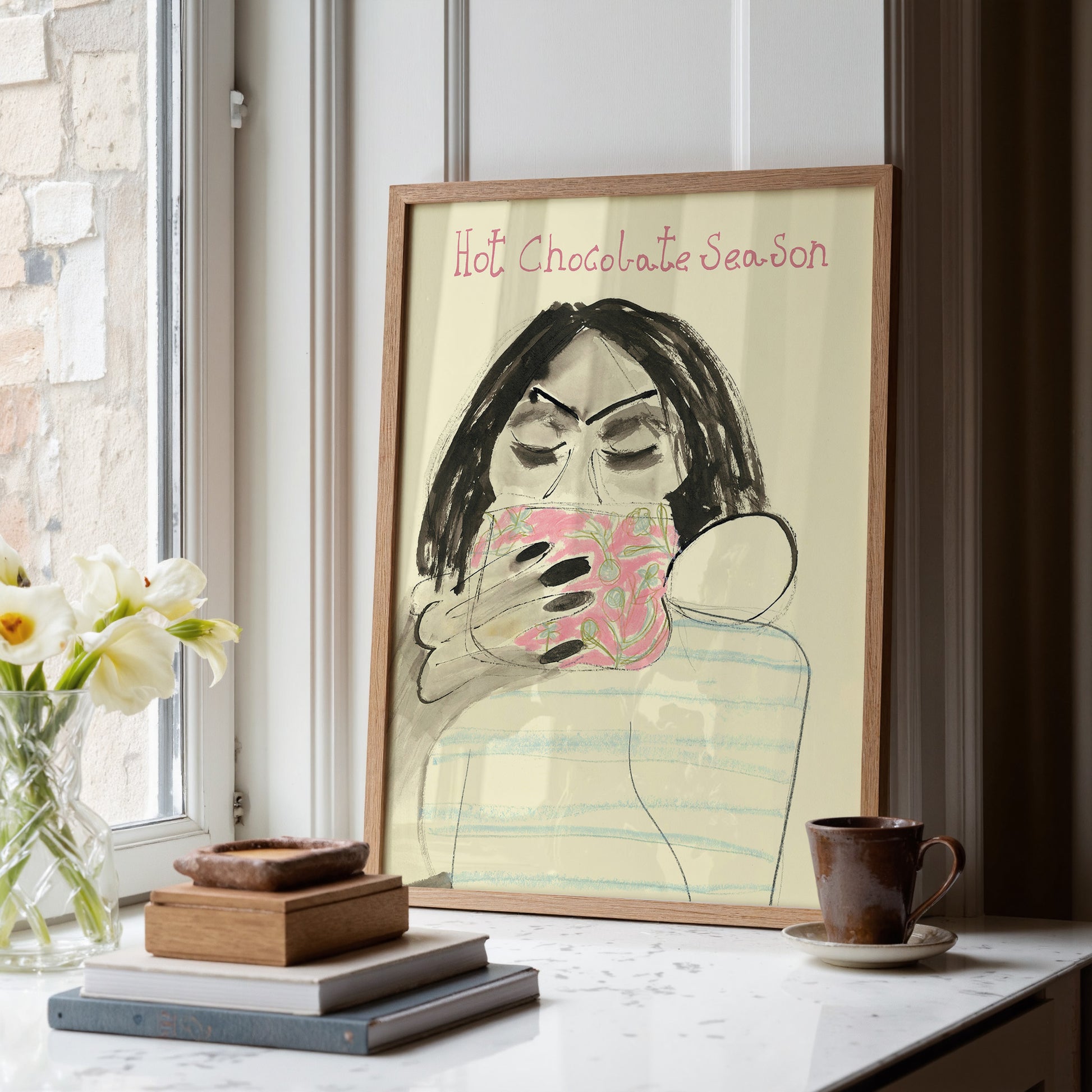 Framed artwork of a person holding a mug with 'Hot Chocolate Season' text, placed on a surface with decor items and a mug.