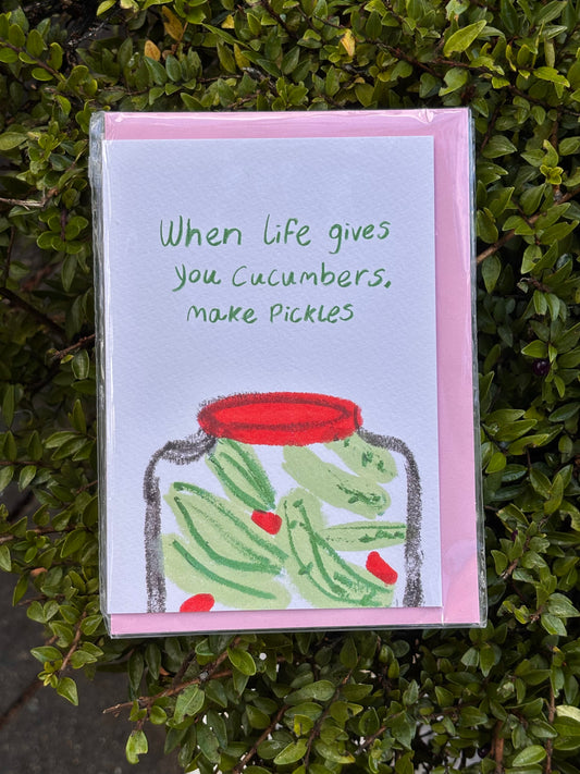 Card with a jar of pickles and text 'When life gives you cucumbers, make pickles' on a green leafy background