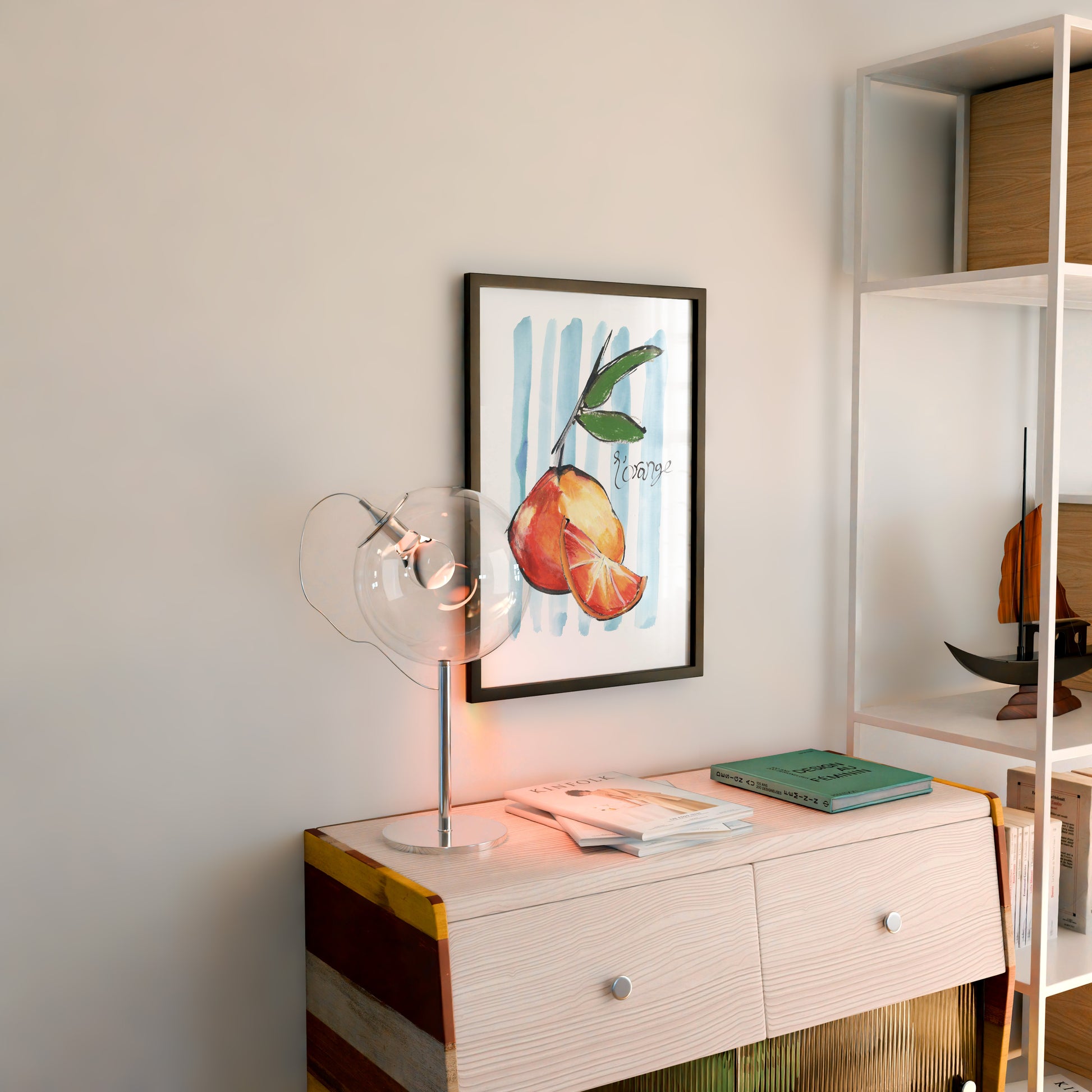 Framed artwork of oranges on a wall above a wooden side table with a lamp and books.