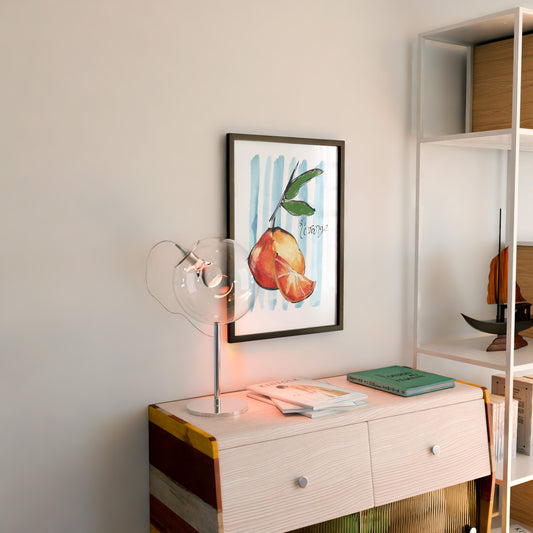 Framed artwork of oranges on a wall above a wooden side table with a lamp and books.
