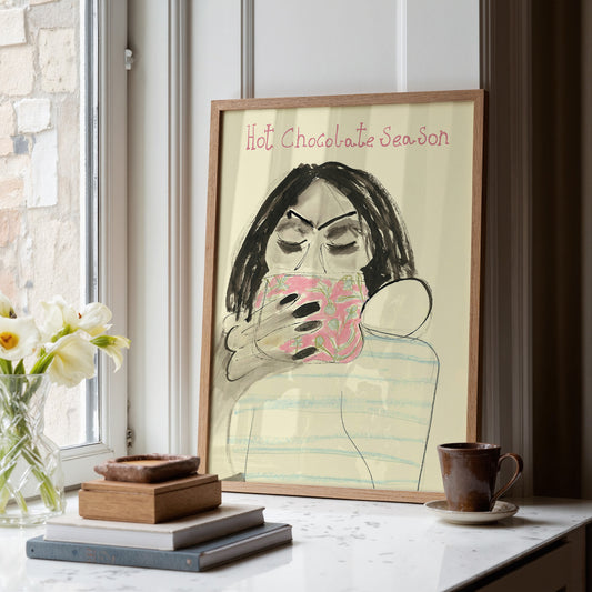 Framed artwork of a person holding a mug with 'Hot Chocolate Season' text, placed on a surface with decor items and a mug.