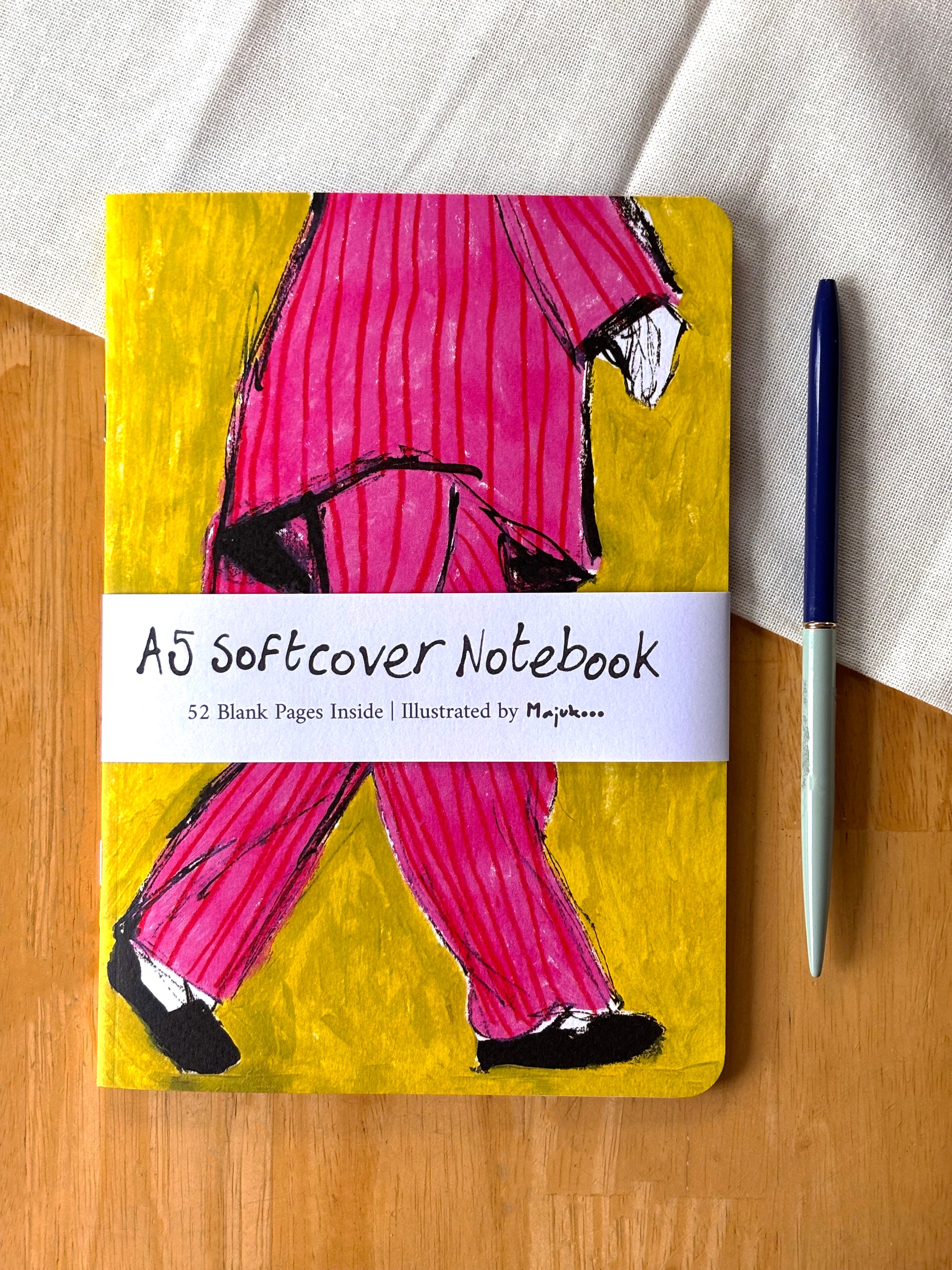 A5 softcover notebook with a colorful cover illustration of a person in pink pants on a wooden surface.