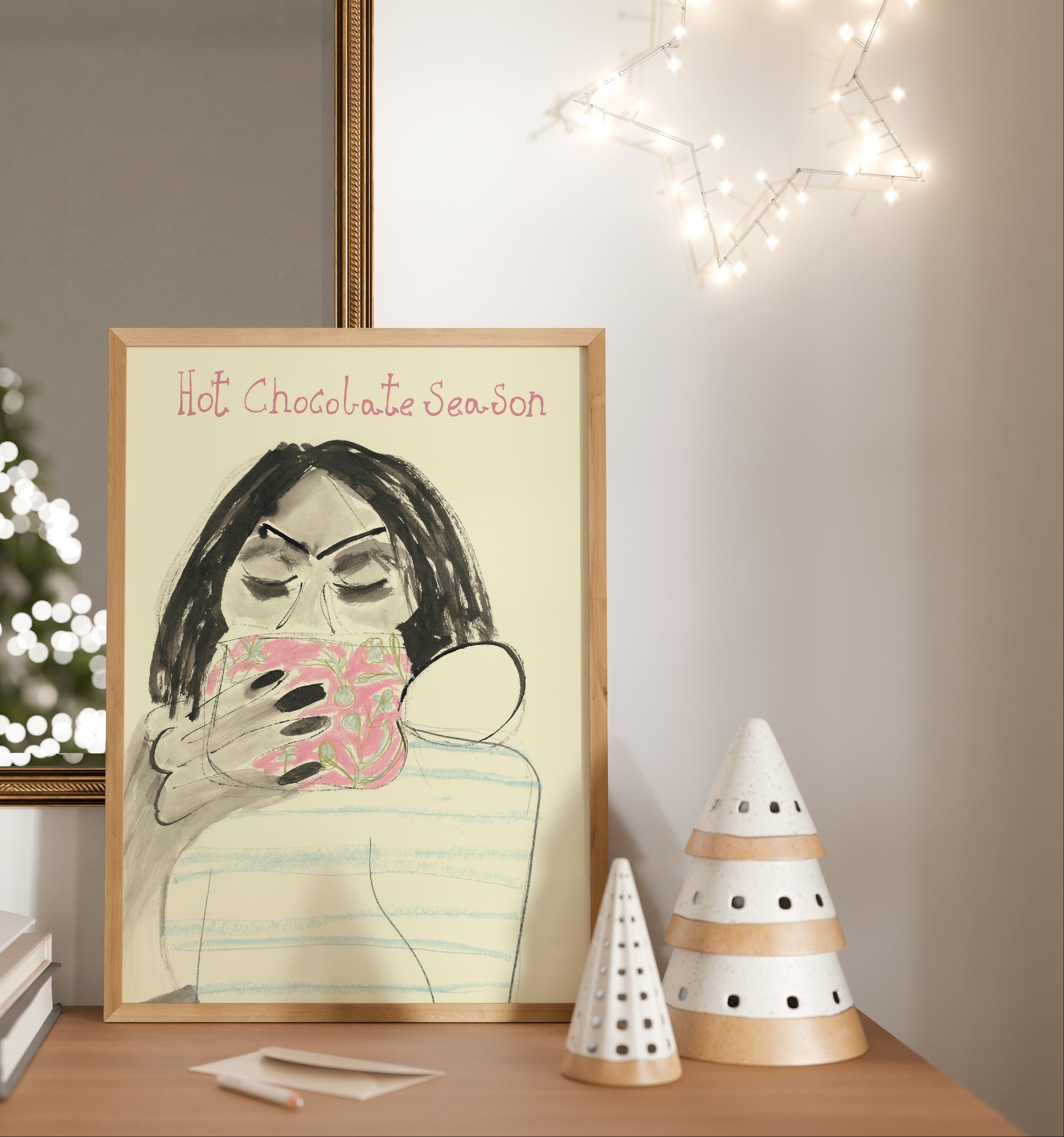 Framed artwork of a person with a hot chocolate mug on a table with decorative lights in the background